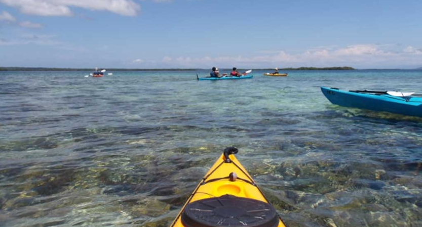 The tip of a yellow kayak juts out over clear, blue water. There are other kayaks on the water in the distance. 